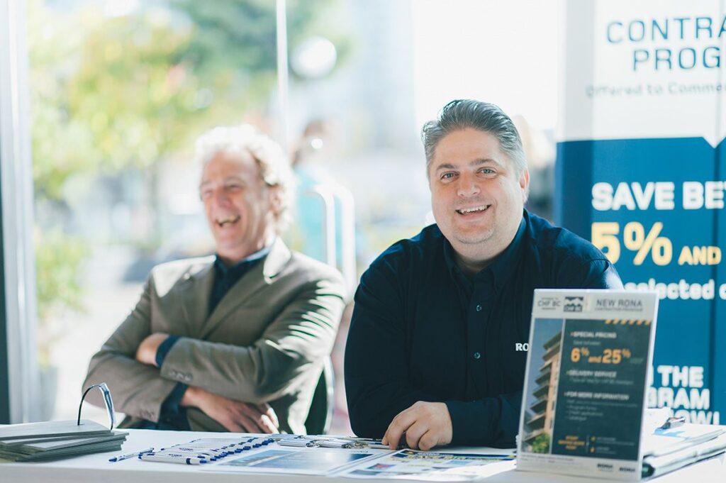 Two men smiling at a Rona vendor table for the group buying program