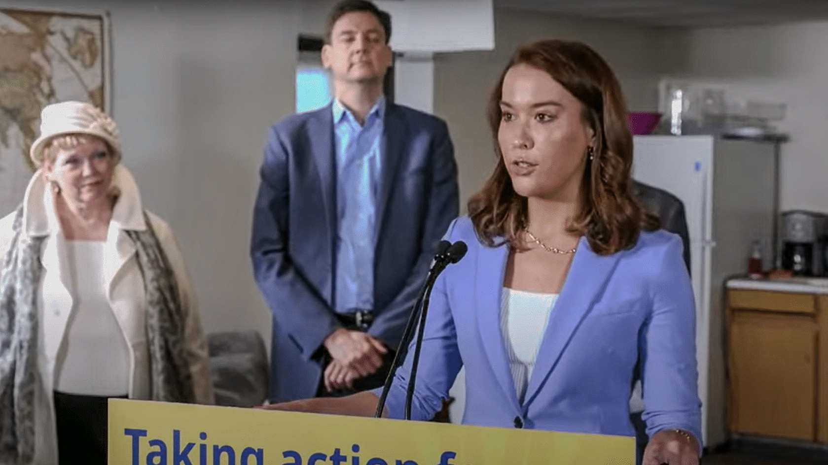 Katie Maslechko speaking at a podium with two people standing behind her